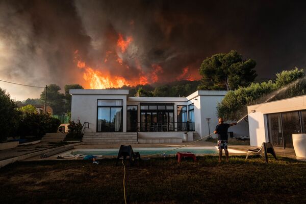 Homem usa mangueira de jardinagem para ensopar sua casa antes de evacuá-la, enquanto incêndio florestal arde por trás de sua residência em La Couronne, França - Sputnik Brasil