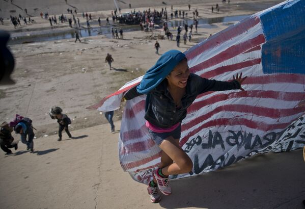 Imigrante ajuda a levar a bandeira norte-americana à fronteira entre o México e os EUA, depois de ter ultrapassado a polícia mexicana na fronteira. A construção de um muro fronteiriço entre os EUA e o México mais fortificado foi uma das promessas da campanha eleitoral de Trump de 2016 - Sputnik Brasil