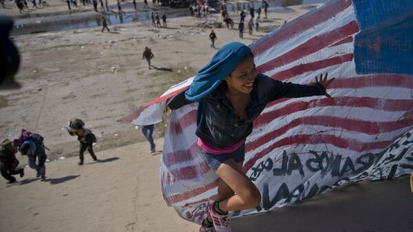 Imigrante ajuda a levar a bandeira norte-americana à fronteira entre o México e os EUA, depois de ter ultrapassado a polícia mexicana na fronteira. A construção de um muro fronteiriço entre os EUA e o México mais fortificado foi uma das promessas da campanha eleitoral de Trump de 2016 - Sputnik Brasil