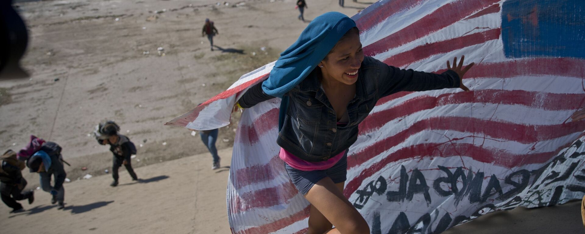 Imigrante ajuda a levar a bandeira norte-americana à fronteira entre o México e os EUA, depois de ter ultrapassado a polícia mexicana na fronteira. A construção de um muro fronteiriço entre os EUA e o México mais fortificado foi uma das promessas da campanha eleitoral de Trump de 2016 - Sputnik Brasil, 1920, 02.11.2024