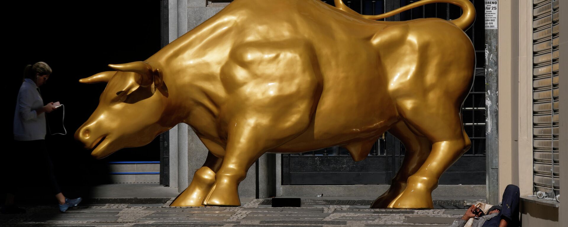 Homem sem-teto dorme perto do Touro de Ouro, réplica do Touro de Wall Street, símbolo do mercado financeiro, fora da B3, São Paulo, 17 de novembro de 2021 - Sputnik Brasil, 1920, 10.04.2025