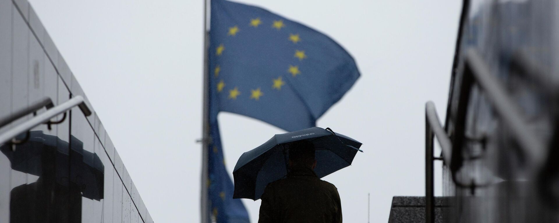 Homem caminha com guarda-chuva perto de bandeiras da União Europeia no exterior da sede do bloco, em Bruxelas, na Bélgica, em 16 de outubro de 2019 (foto de arquivo) - Sputnik Brasil, 1920, 08.09.2022