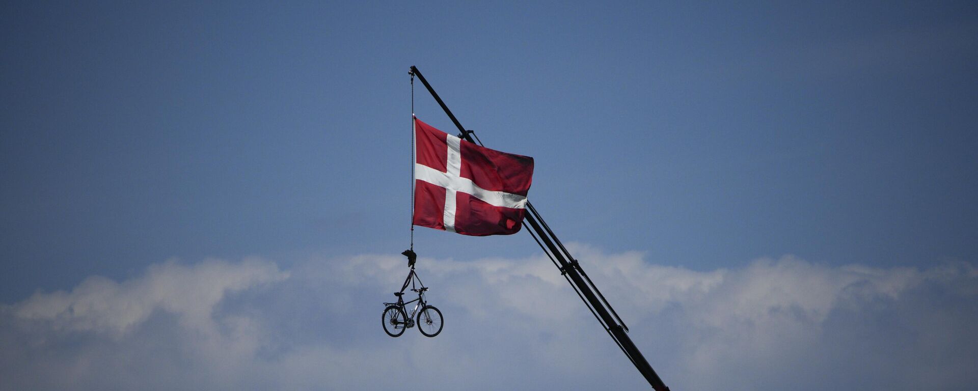 Bandeira dinamarquesa hasteada e uma bicicleta está suspensa de um guindaste enquanto os ciclistas passam durante a segunda etapa da corrida de bicicleta Tour de France, no sábado, 2 de julho de 2022. - Sputnik Brasil, 1920, 15.03.2023