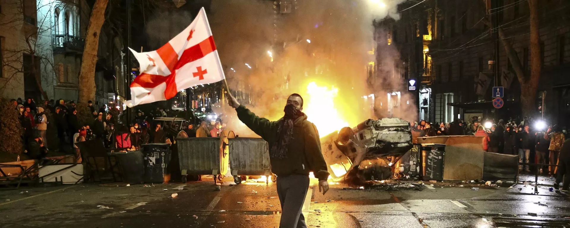 Um homem acena com uma bandeira nacional da Geórgia em frente a uma barricada em chamas enquanto outros manifestantes ficam atrás, não muito longe do edifício do parlamento georgiano em Tbilisi, Geórgia, 9 de março de 2023. - Sputnik Brasil, 1920, 08.05.2024