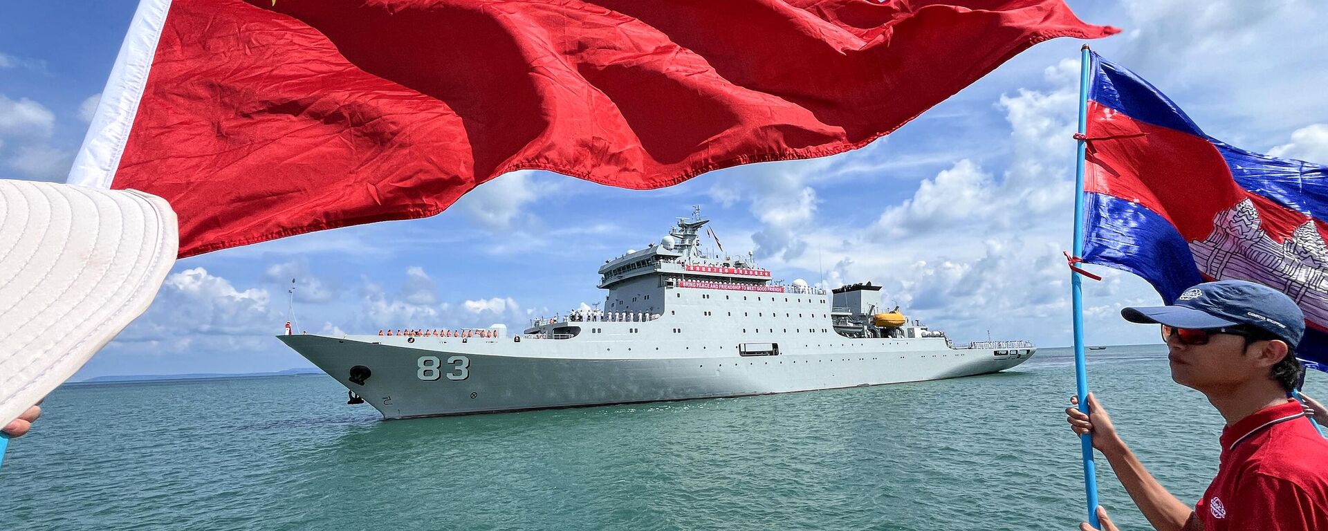 Pessoas agitam bandeiras do Camboja (R) e da China (L) enquanto o navio-escola chinês Qijiguang se prepara para atracar com uma faixa que diz Traga paz e amizade para encontrar bons amigos durante uma cerimônia de boas-vindas no porto de Sihanoukville, na província de Preah Sihanouk, em 19 de maio de 2024 - Sputnik Brasil, 1920, 25.05.2024