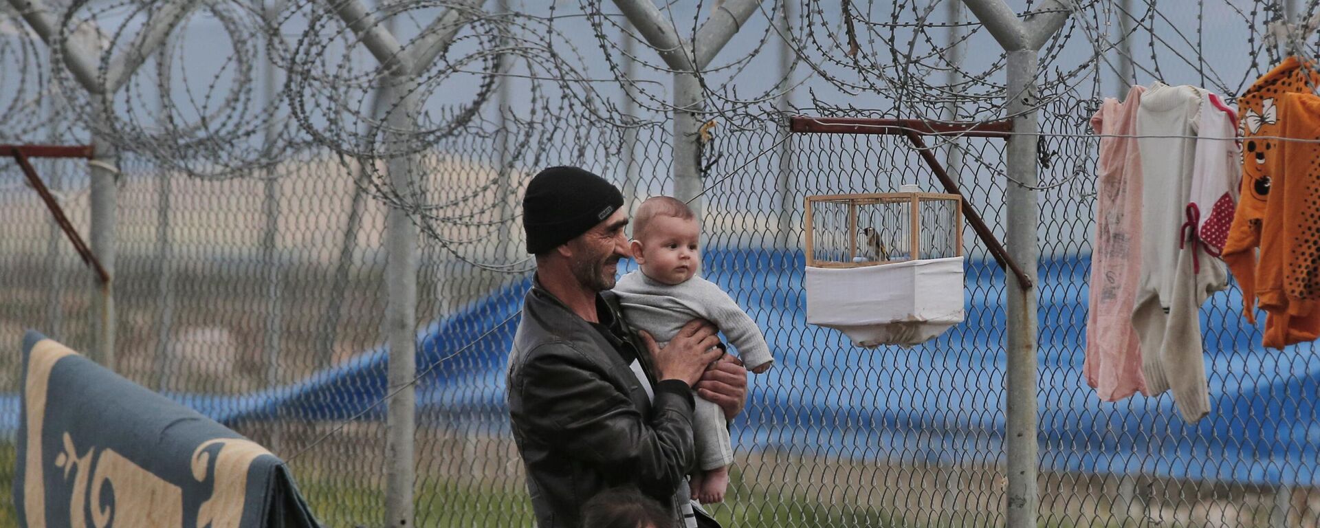 Refugiado sírio em um campo para refugiados em Islahiye, na Turquia, com o filho de 6 meses e a filha de 6 anos, observa o pássaro de estimação da família, suspenso em uma gaiola na cerca de arame farpado do campo, em 16 de março de 2016 - Sputnik Brasil, 1920, 04.12.2024