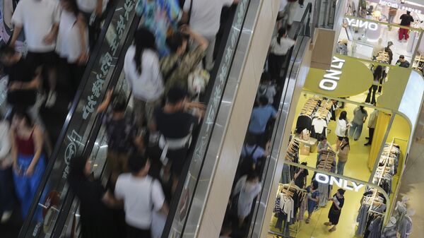 Pessoas sobem escadas rolantes enquanto compradores, à direita no andar de baixo, selecionam roupas em uma loja dentro de um shopping em Pequim, 13 de julho de 2025 - Sputnik Brasil