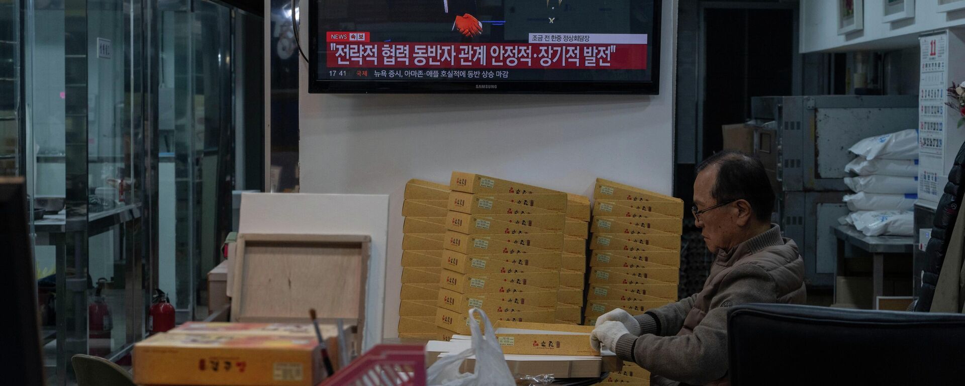 Um homem empacotando produtos em uma padaria enquanto uma televisão mostra o presidente chinês Xi Jinping (E) e o presidente sul-coreano Lee Jae Myung apertando as mãos durante uma reunião bilateral à margem da Cúpula da Cooperação Econômica Ásia-Pacífico (APEC) de 2025, realizada em Gyeongju, Coreia do Sul, 1º de novembro de 2025 - Sputnik Brasil, 1920, 07.01.2026