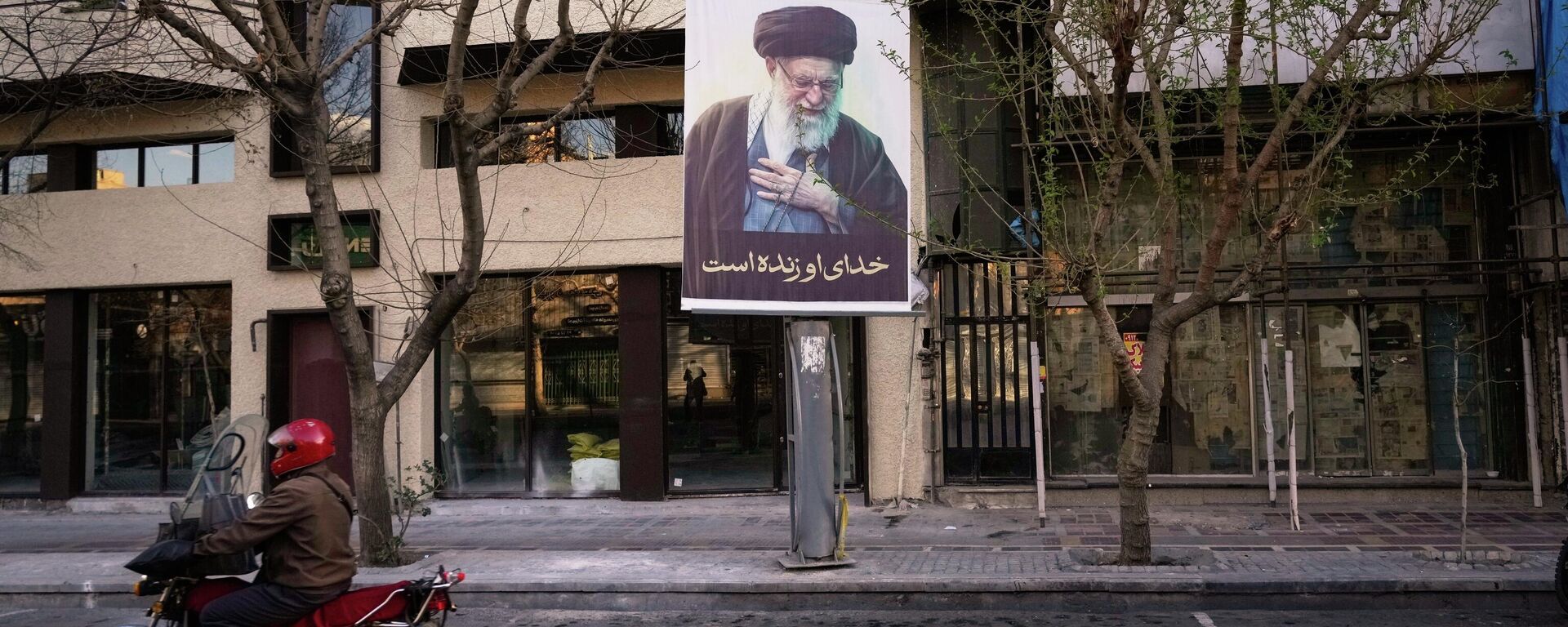 Uma motocicleta passa por uma foto do falecido Líder Supremo iraniano, Aiatolá Ali Khamenei, em uma rua vazia de Teerã, Irã, no domingo, 1º de março de 2026, após a confirmação da morte de Khamenei em ataques dos EUA e de Israel. - Sputnik Brasil, 1920, 01.03.2026