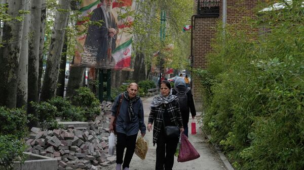 Mulheres caminham ao lado de uma faixa que retrata o aiatolá Ali Khamenei, morto nos ataques dos EUA e de Israel em 28 de fevereiro, no norte de Teerã, Irã. 12 de abril de 2026 - Sputnik Brasil