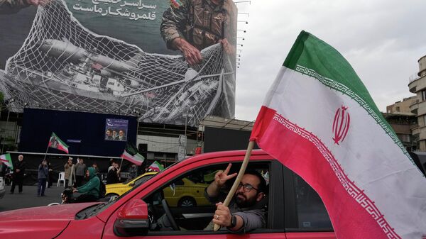 Homem faz o sinal de vitória enquanto carrega bandeira iraniana em frente a um cartaz anti-EUA, em Teerã, que retrata aeronaves norte-americanas caindo em uma rede de pesca das Forças Armadas iranianas. Irã, 13 de abril de 2026 - Sputnik Brasil