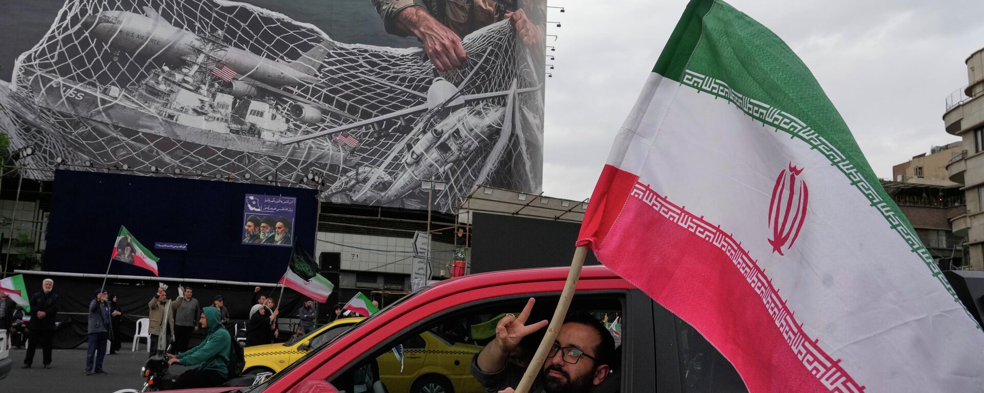 Homem faz o sinal de vitória enquanto carrega bandeira iraniana em frente a um cartaz anti-EUA, em Teerã, que retrata aeronaves norte-americanas caindo em uma rede de pesca das Forças Armadas iranianas. Irã, 13 de abril de 2026 - Sputnik Brasil, 1920, 21.04.2026