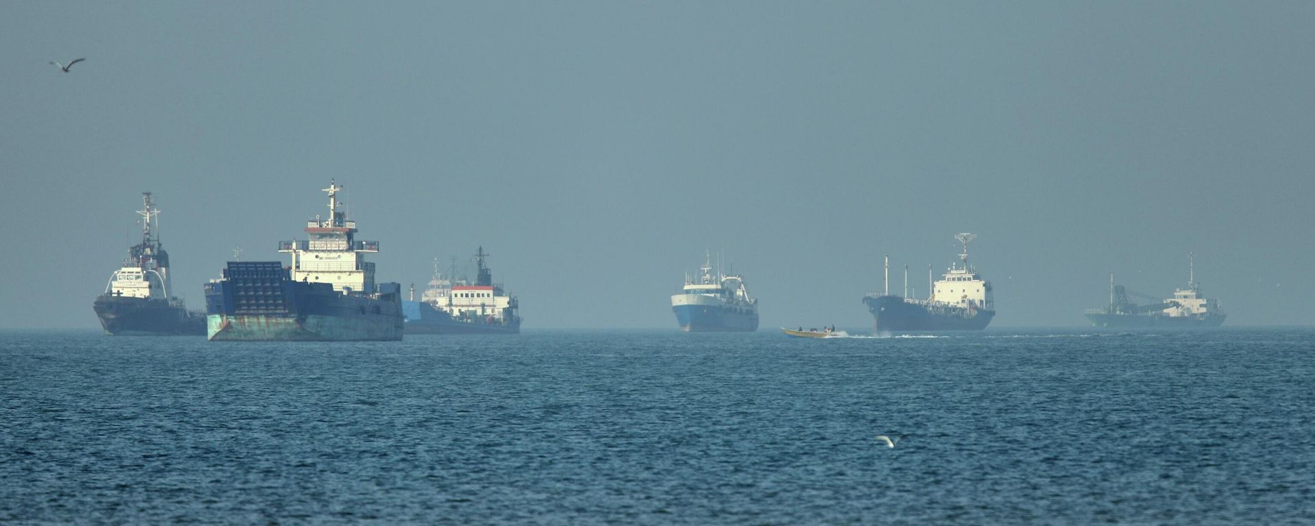 Navios de carga, incluindo graneleiros e navios de carga geral, estão ancorados ao largo da costa, enquanto uma pequena lancha passa em primeiro plano, no estreito de Ormuz, perto de Bandar Abbas. Irã, 4 de maio de 2026 - Sputnik Brasil, 1920, 26.06.2026