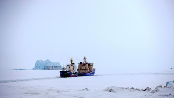 Um quebra-gelo abrindo caminho para um navio cargueiro com um iceberg ao fundo, próximo a um porto na ilha de Alexandra Land, perto de Nagurskoye, Rússia, 17 de maio de 2021 - Sputnik Brasil
