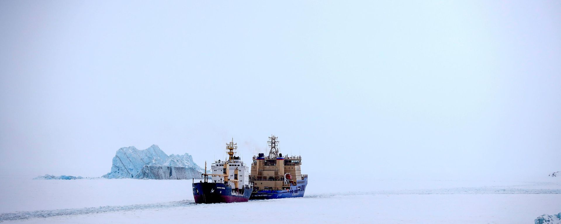 Um quebra-gelo abrindo caminho para um navio cargueiro com um iceberg ao fundo, próximo a um porto na ilha de Alexandra Land, perto de Nagurskoye, Rússia, 17 de maio de 2021 - Sputnik Brasil, 1920, 10.08.2026