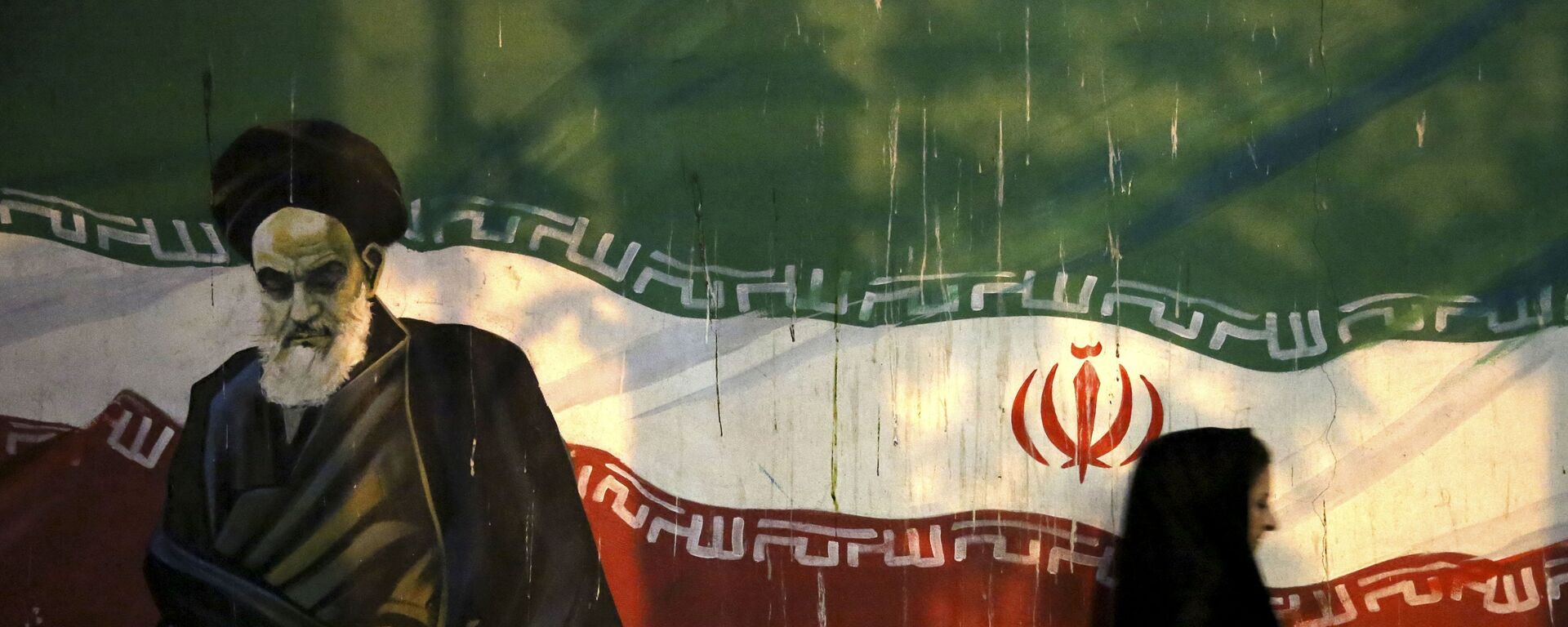 Iraniana coberta passa por muro que retrata o revolucionário aiatolá Ruhollah Khomeini e a bandeira iraniana nacional, pintada na parede da antiga Embaixada dos EUA, em Teerã, no Irã - Sputnik Brasil, 1920, 19.04.2024