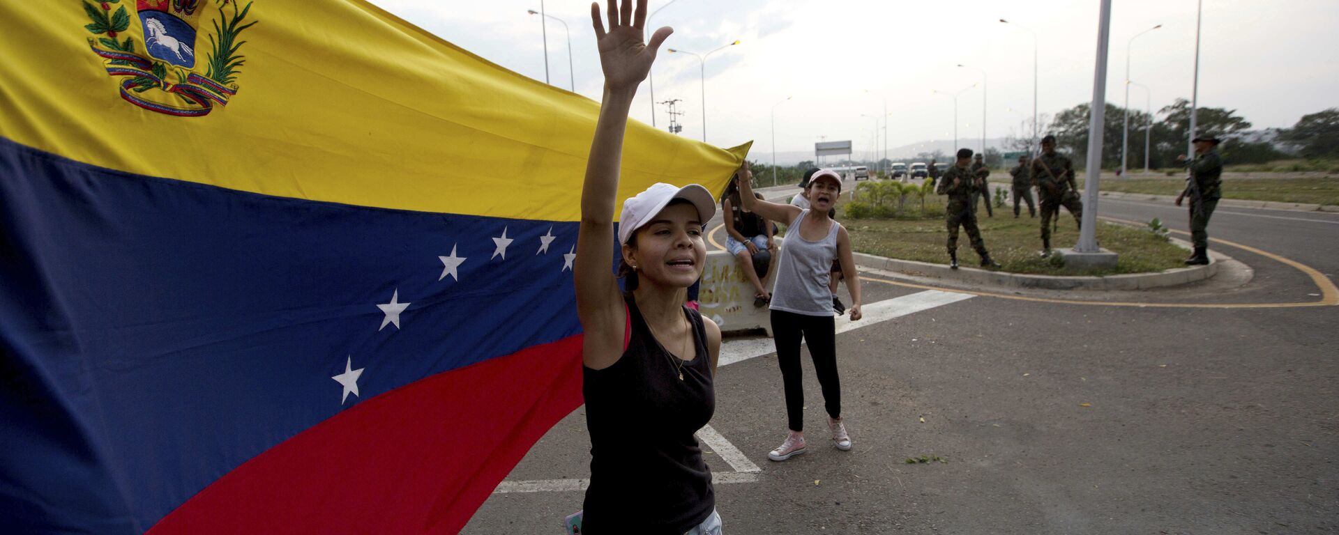 Mulher grita bem-vinda ajuda humanitária, balançando bandeira venezuelana em frente a grupo de soldados do Exército venezuelano e oficiais da Guarda Nacional bloqueando o acesso principal à ponte internacional Tienditas, que liga Colômbia e Venezuela, 7 de fevereiro de 2019 - Sputnik Brasil, 1920, 29.07.2024